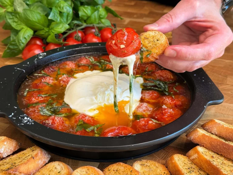 Geroosterde tomaten geserveerd met burrata en baguette op een houten snijplank